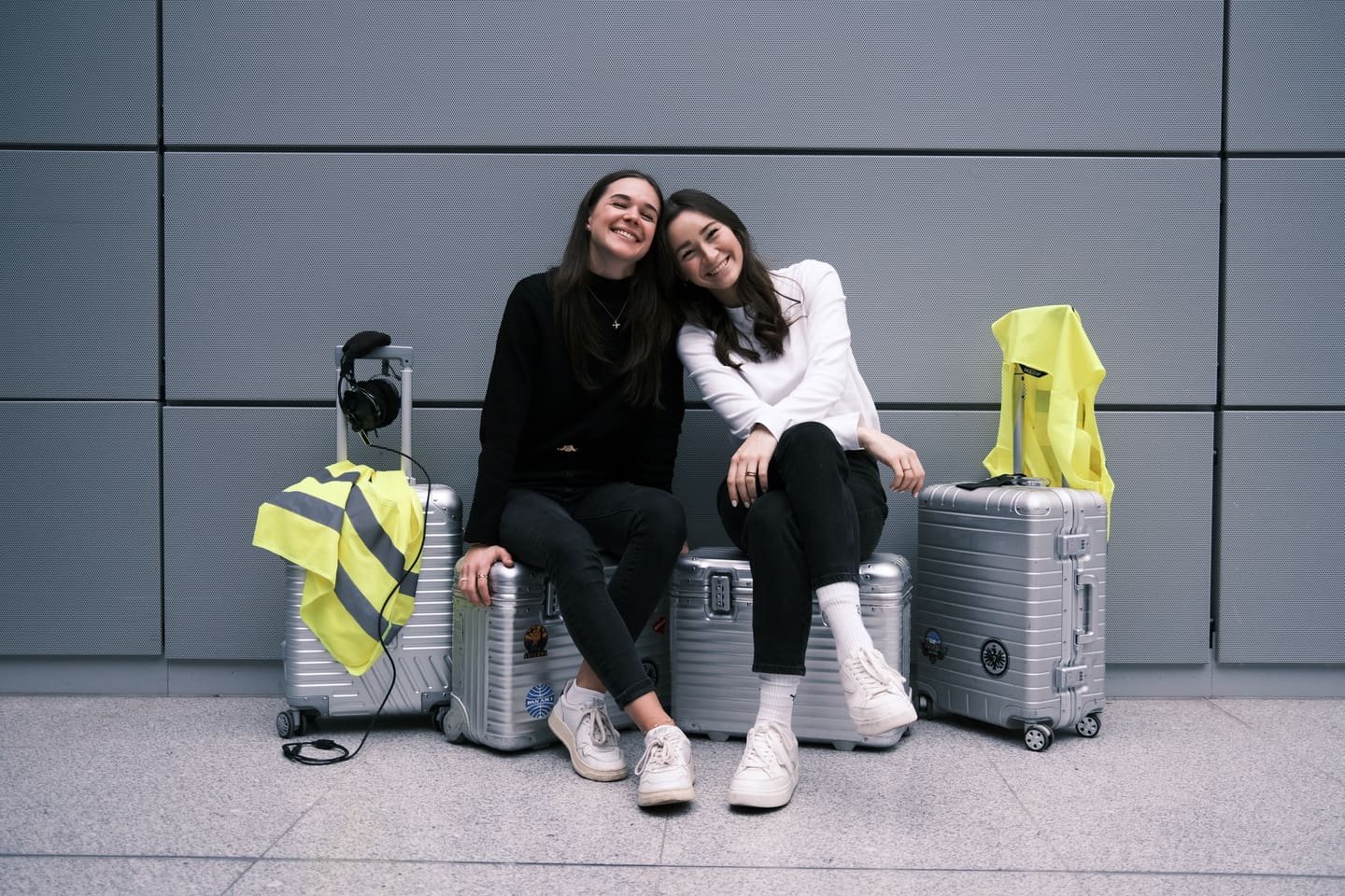 Anna und Julia sitzen lachend auf silbernen Koffern vor einer grauen Wand, daneben Warnwesten und ein Headset.